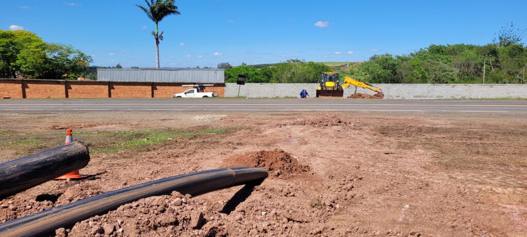 A imagem mostra uma área de construção em um dia ensolarado. No primeiro plano, há tubos pretos de grande diâmetro parcialmente enterrados em solo avermelhado. Um cone de trânsito laranja está posicionado próximo a um dos tubos. Ao fundo, uma estrada divide a cena. Do lado esquerdo da estrada, há um muro de tijolos vermelhos com uma estrutura longa e cinza atrás dele, possivelmente um contêiner. Uma palmeira alta se destaca contra o céu azul. Um veículo branco, possivelmente uma caminhonete, está estacionado próximo ao muro. Do lado direito da estrada, um muro de blocos de concreto cinza se estende. Uma escavadeira amarela está em operação, com seu braço mecânico cavando perto do muro. Uma pessoa vestindo roupas azuis está próxima à escavadeira. A paisagem ao fundo é composta por vegetação verde e colinas suaves sob um céu azul claro com algumas nuvens brancas.
