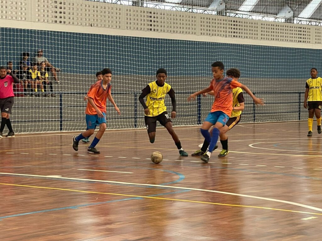 A imagem mostra um jogo de futebol de salão entre jovens jogadores em uma quadra interna. Os jogadores vestem uniformes de cores vibrantes, com destaque para os laranjas e amarelos. A quadra possui piso de madeira polida e linhas brancas e amarelas demarcando o espaço de jogo. Ao fundo, uma rede azul separa a quadra da área onde espectadores estão sentados em arquibancadas. A iluminação é artificial, típica de ambientes esportivos fechados, e a atmosfera parece ser de um jogo dinâmico e competitivo.