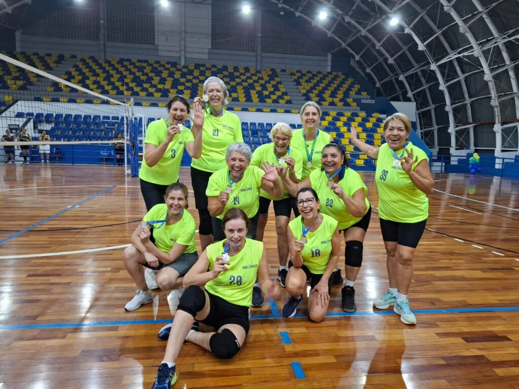 A imagem mostra um grupo de mulheres em uma quadra de vôlei, todas vestindo uniformes verde-limão e exibindo medalhas. Elas parecem estar comemorando uma vitória ou um evento. Algumas mulheres estão agachadas na frente, enquanto outras estão em pé atrás delas. Todas sorriem e parecem felizes, com algumas levantando suas medalhas. Ao fundo, há arquibancadas azuis e amarelas e uma rede de vôlei. A iluminação sugere que a foto foi tirada em um ginásio.