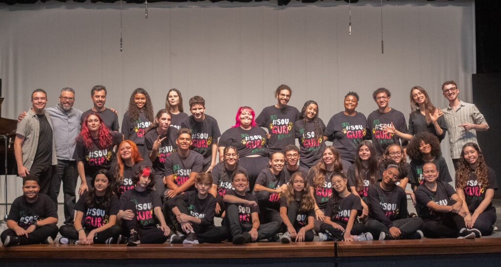 A imagem mostra um grupo grande de pessoas, predominantemente jovens, posando para uma foto em um palco. Eles estão dispostos em várias fileiras, com alguns sentados na frente e outros em pé atrás. A maioria das pessoas está vestindo camisetas pretas com a inscrição "#SOU GURP MÚSICA" em cores vibrantes como rosa, amarelo e verde. Alguns adultos estão presentes no grupo, aparentemente liderando ou acompanhando os jovens. Um homem com óculos e camisa cinza está no meio da fileira de trás, ao lado de outro homem com camisa preta e um terceiro com uma camisa listrada clara. Uma mulher com cabelo comprido e escuro, vestindo uma camisa preta, está na fileira do meio, acenando para a câmera. O fundo é um palco com cortinas e alguns equipamentos de iluminação ou som pendurados. A iluminação sugere que a foto foi tirada em um ambiente de apresentação. As expressões faciais variam de sorrisos a olhares mais sérios, mas a atmosfera geral parece ser de união e talvez celebração de um evento musical ou artístico.