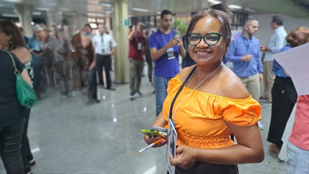 A fotografia mostra uma mulher sorridente, usando óculos verdes e uma blusa laranja de ombros à mostra, em um evento. Ela segura dois celulares e uma caneta na mão direita. Ao fundo, outras pessoas em um ambiente que parece ser um corredor ou salão de eventos, com algumas delas usando crachás. A iluminação é clara e o foco principal está na mulher à frente.