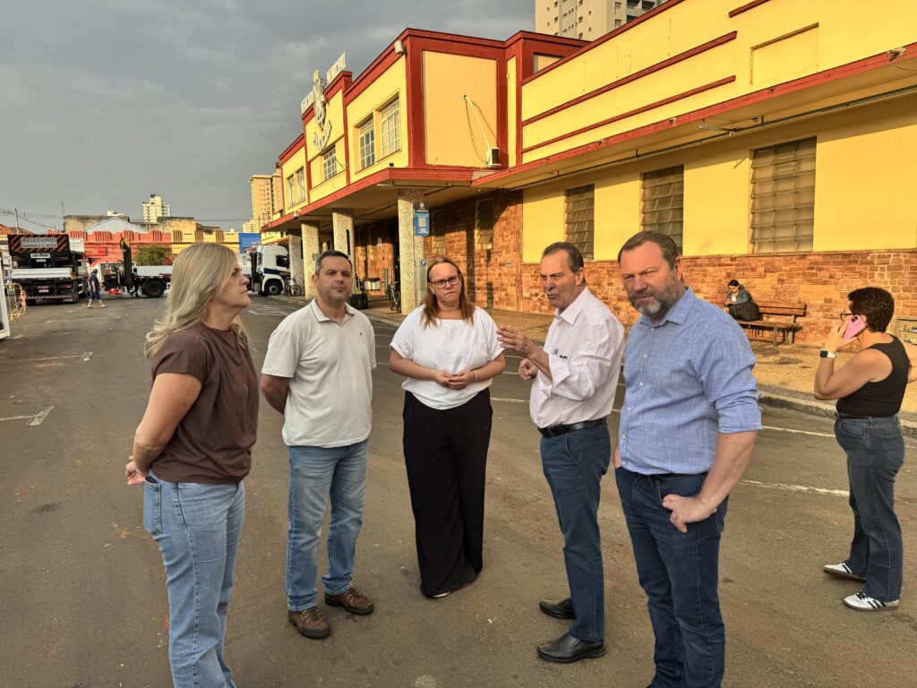 Um grupo de cinco pessoas está em pé em um estacionamento. Um edifício com arquitetura amarela e vermelha está ao fundo, com algumas janelas de vidro fosco. O céu está nublado. Da esquerda para a direita, as pessoas são: * Uma mulher com cabelo loiro comprido, vestindo uma camiseta marrom e jeans, está de pé com as mãos juntas atrás das costas. * Um homem com cabelo escuro curto, vestindo uma camisa polo bege e jeans, está de pé com as mãos juntas atrás das costas. * Uma mulher com cabelo castanho na altura do queixo, vestindo uma camiseta branca e calças pretas largas, está de pé com as mãos cruzadas. * Um homem com cabelo escuro curto e grisalho, vestindo uma camisa rosa clara e jeans, está gesticulando enquanto fala. * Um homem com cabelo castanho curto e barba, vestindo uma camisa azul e jeans, está de pé com a mão no bolso.