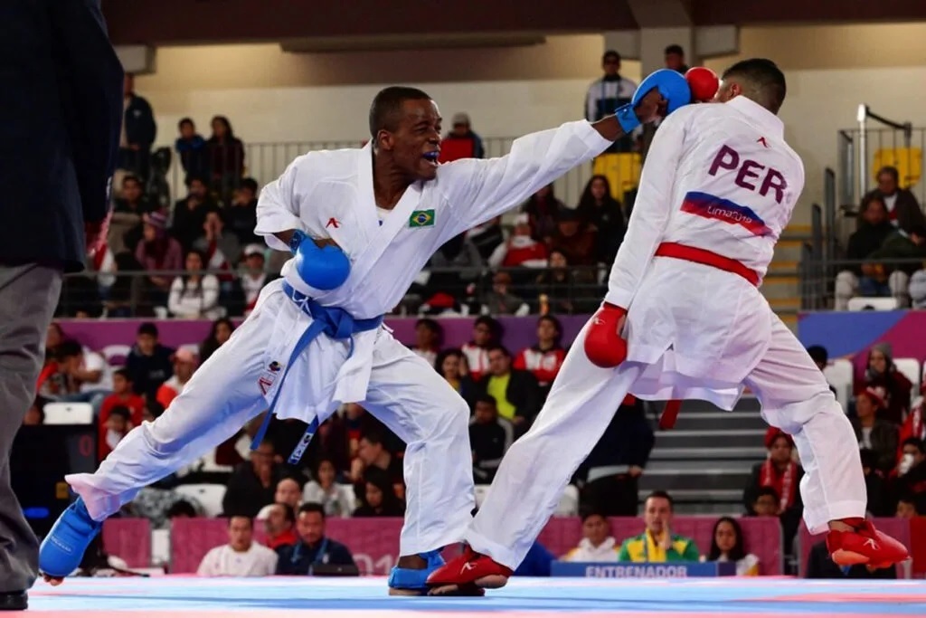 agem mostra dois atletas de karatê competindo em um tatame. O atleta da esquerda, vestindo um quimono branco com faixa azul e luvas azuis, está desferindo um golpe de soco com a mão direita. Ele tem a bandeira do Brasil em seu quimono. O atleta da direita, vestindo um quimono branco com faixa vermelha e luvas vermelhas, está de costas para o espectador e parece estar se defendendo ou esquivando do golpe. Seu quimono exibe a sigla "PER", indicando que ele representa o Peru. Ambos os atletas estão em posições dinâmicas, com as pernas afastadas e em movimento, demonstrando a intensidade da luta. Ao fundo, uma plateia assiste à competição em arquibancadas.