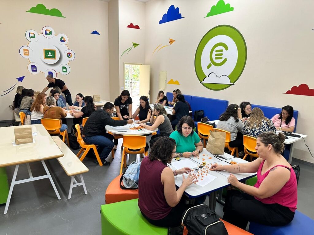 A imagem mostra um grupo de pessoas, a maioria mulheres, reunidas em uma sala clara e colorida, possivelmente uma sala de aula ou um espaço de workshop. Elas estão sentadas em mesas redondas e retangulares, engajadas em uma atividade que parece envolver a montagem de estruturas com pequenas peças coloridas. Há um instrutor ou facilitador, um homem, em pé no fundo, orientando alguns participantes. As paredes da sala são decoradas com elementos lúdicos e educativos, como nuvens coloridas, aviões de papel e um grande círculo verde com um símbolo de euro estilizado e uma lupa, sugerindo um tema relacionado à criatividade, aprendizado ou inovação. A iluminação é boa, vindo de cima e de uma janela visível ao fundo, criando um ambiente acolhedor e propício para atividades em grupo. Há também móveis coloridos, como cadeiras laranjas e bancos em tons de verde e laranja, que adicionam vivacidade ao espaço.