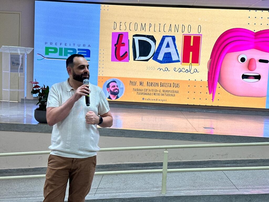 A imagem mostra um homem barbudo, vestindo uma camisa polo bege e calças marrons, segurando um microfone e falando em um palco. Atrás dele, um grande telão exibe uma apresentação. O telão tem o logotipo da "PREFEITURA PIRA" à esquerda. O restante do telão é dominado por um título em letras grandes e coloridas que diz "DESCOMPLICANDO O TDAH na escola", com uma ilustração estilizada de um rosto com cabelo rosa vibrante à direita. Abaixo do título principal, há um círculo com a foto do palestrante e seu nome, "PROF. ME. ROBSON BATISTA DIAS", seguido por suas qualificações: "PSICÓLOGO (CRP 14/05584-6), NEUROPSICÓLOGO, PSICOPEDAGOGO E MESTRE EM PSICOLOGIA". Abaixo de suas credenciais, está seu nome de usuário no Instagram: "@robsondiaspsi". Há também um vaso com plantas vermelhas no canto esquerdo do palco.