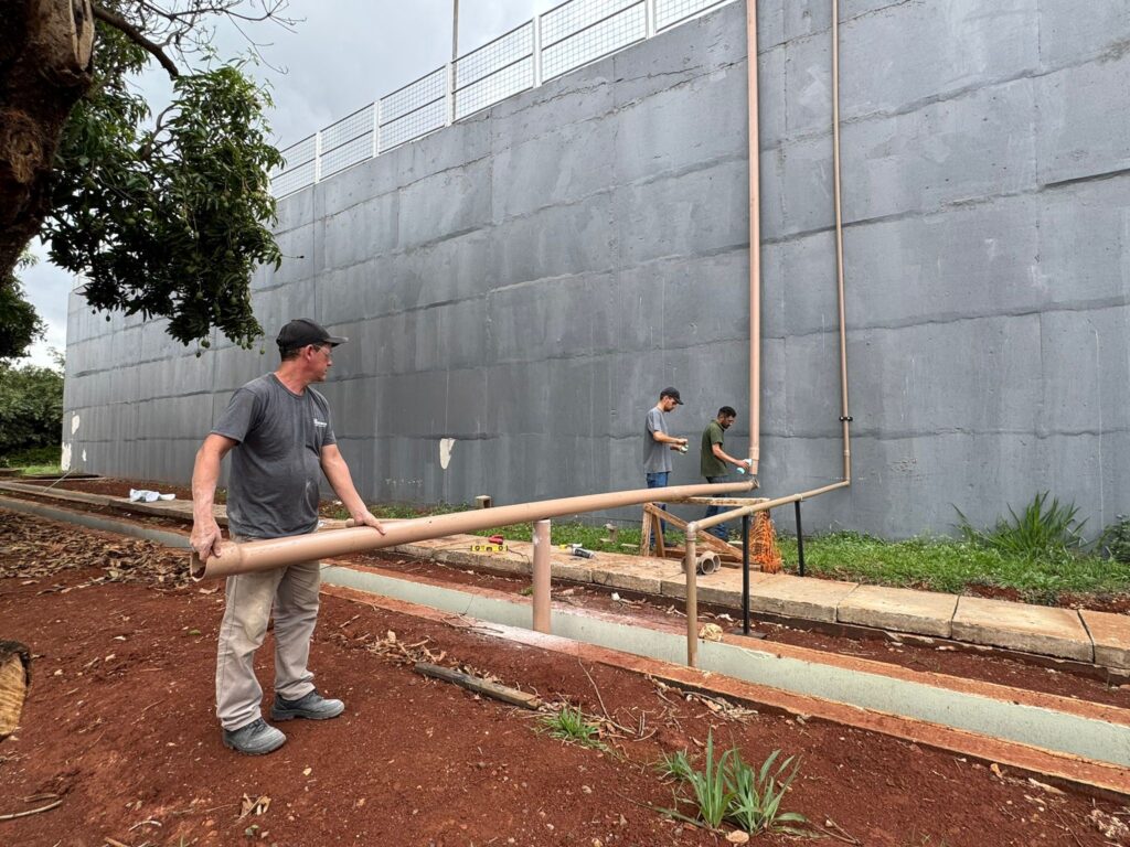 A imagem mostra três homens trabalhando na instalação de tubulações de PVC de cor bege, em frente a um grande muro de concreto cinza. O homem mais próximo, na frente esquerda, está segurando um longo pedaço de tubo horizontalmente, olhando para a direita. Ele usa uma camiseta cinza, calça bege e um boné preto. Dois outros homens estão mais atrás, perto do muro. Um deles, com uma camiseta cinza e boné preto, parece estar segurando uma lata em spray. O outro homem, vestindo uma camiseta verde, também parece estar usando uma lata em spray em uma tubulação vertical. Ambos estão em pé sobre uma calçada de concreto, com grama crescendo ao redor. O muro de concreto é alto e tem uma textura áspera, com algumas marcas de reparo visíveis. No topo do muro, há uma grade de metal com uma cerca de arame. À esquerda da imagem, um ramo de uma árvore com folhas verdes e alguns frutos (possivelmente mangas) se estende sobre a cena. O solo em primeiro plano é de cor marrom-avermelhada, com algumas folhas secas e pequenos brotos de grama. Há também uma vala ou canaleta visível no solo, com algumas tubulações menores correndo ao longo dela.