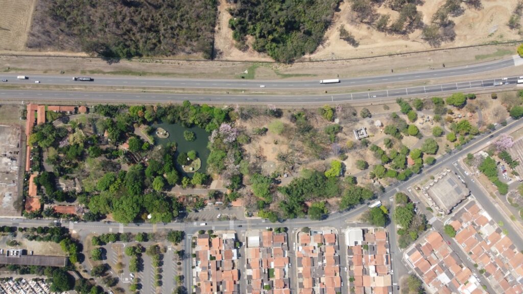 A imagem aérea revela uma paisagem urbana e natural entrelaçadas. Uma rodovia movimentada corta a cena horizontalmente, ladeada por áreas verdes e edifícios. À esquerda da rodovia, observa-se um parque com um lago sereno, pontilhado por pequenas ilhas. Árvores frondosas e algumas flores rosadas criam uma atmosfera bucólica. Ao lado do parque, há um complexo de edifícios com telhados alaranjados, possivelmente residências ou instalações administrativas. À direita da rodovia, a paisagem se transforma em áreas mais secas com vegetação esparsa e algumas árvores isoladas. Mais adiante, uma área residencial com telhados de barro se estende, marcada por ruas e estacionamentos. Um grande edifício com painéis solares no telhado também é visível, sugerindo uma estrutura com foco em sustentabilidade. A imagem captura a diversidade de ambientes, desde a infraestrutura rodoviária até áreas de lazer e moradia.