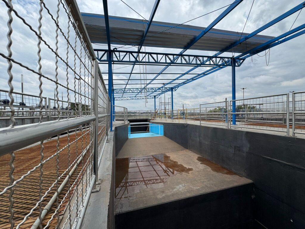 A imagem mostra uma estrutura industrial com um telhado de metal azul e cinza, sustentado por vigas azuis. Há uma grade de metal na frente, com um corrimão de aço inoxidável. Abaixo da grade, há uma área com uma superfície de metal enferrujado. No centro da imagem, há uma piscina ou tanque com água azul e uma área de concreto ao redor, que tem algumas poças de água refletindo a estrutura do telhado. Ao fundo, pode-se ver mais cercas de metal, equipamentos industriais e um céu nublado. A imagem parece ter sido tirada de um ponto de vista ligeiramente elevado, olhando para dentro da estrutura.