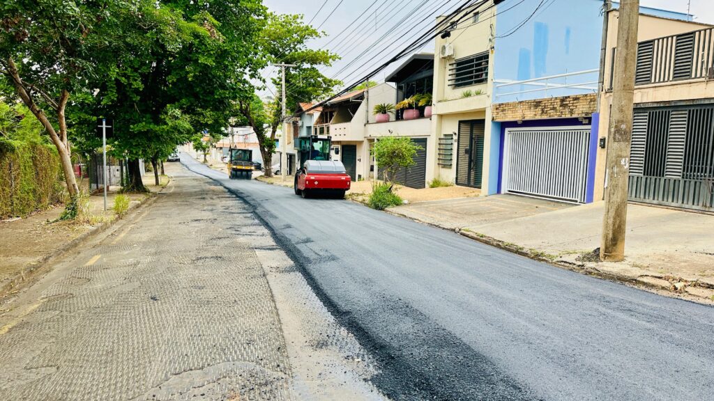 A imagem mostra uma rua residencial em processo de recapeamento asfáltico. * No primeiro plano, vemos o asfalto recém-aplicado, brilhante e escuro, contrastando com o pavimento antigo e desgastado à esquerda. * Uma máquina de rolo compactador vermelha está em movimento, alisando o novo asfalto. * Mais adiante na rua, outra máquina, um rolo compactador amarelo, também está trabalhando. * As casas à direita exibem arquitetura moderna, com portões de garagem brancos e detalhes em azul e bege. * Árvores frondosas e a vegetação nas calçadas adicionam um toque de verde à cena. * O céu está nublado, sugerindo um dia de trabalho contínuo na pavimentação da rua.