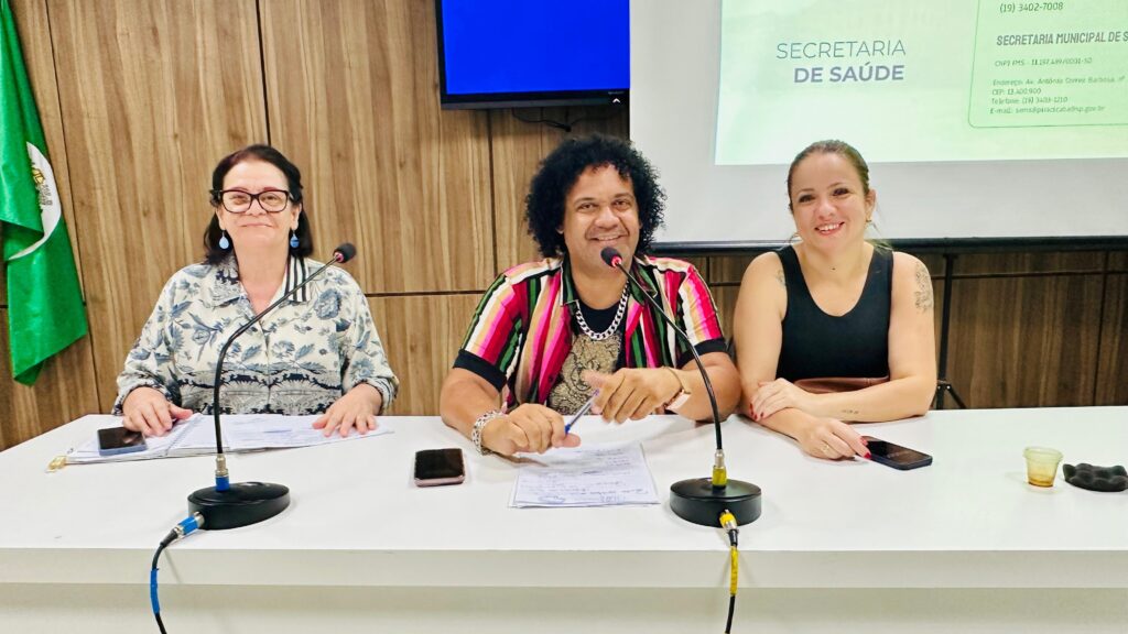 A imagem mostra três pessoas sentadas em uma mesa, com microfones à frente delas. À esquerda, uma mulher de óculos e blusa estampada sorri para a câmera. Ao centro, um homem com cabelo crespo e uma camisa colorida e vibrante está com uma caneta na mão. À direita, uma mulher com um coque e blusa preta também sorri. Atrás deles, um banner com o texto "SECRETARIA DE SAÚDE" é visível, juntamente com uma bandeira verde à esquerda e uma tela de computador com fundo azul no canto superior esquerdo. A mesa parece estar em uma sala de reuniões ou auditório, com uma parede de madeira ao fundo.