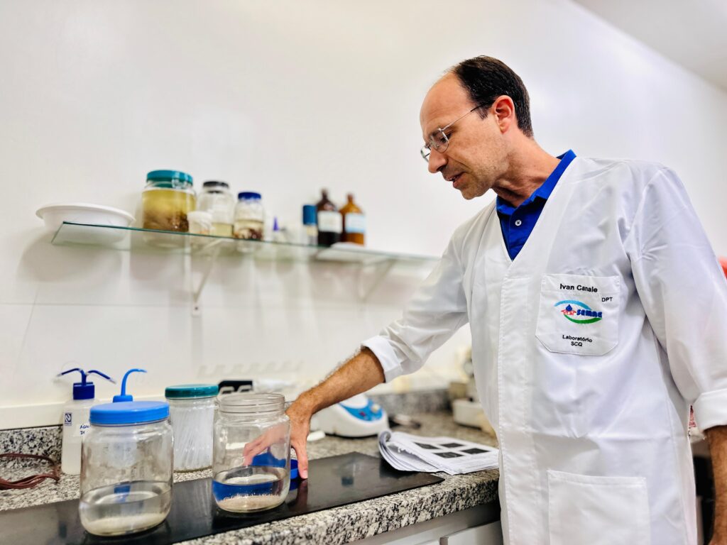 Nesta imagem, um cientista de jaleco branco está em um laboratório. Ele usa óculos e tem uma expressão concentrada enquanto olha para um frasco de vidro em uma bancada. A bancada possui outros recipientes, incluindo um frasco com líquido amarelado e outros com amostras. Ao fundo, uma prateleira exibe mais frascos e garrafas, alguns com líquidos e sólidos. O jaleco do cientista tem um bordado com o nome "Ivan Canale", "DPT", e o logotipo de uma instituição com as palavras "Laboratório SCQ". A iluminação sugere um ambiente de trabalho organizado e funcional.