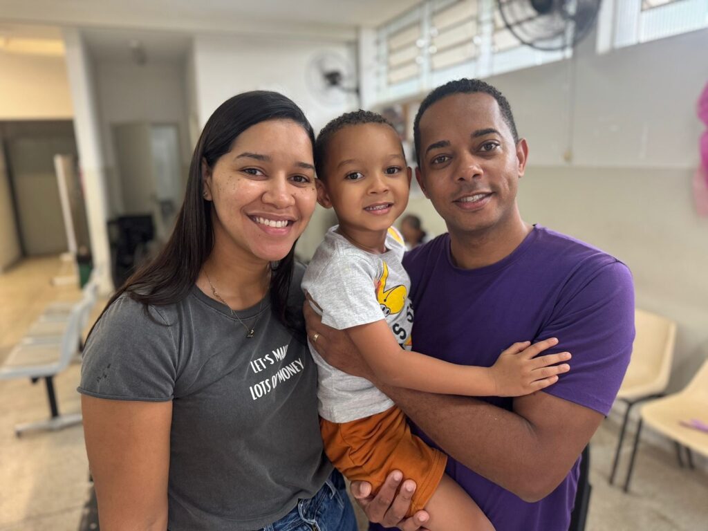 A imagem mostra uma família feliz posando para uma foto. No centro, um menino pequeno, com pele morena e cabelo curto e encaracolado, está sendo abraçado pelo seu pai, que tem pele morena e veste uma camiseta roxa. O menino usa uma camiseta cinza com um desenho amarelo e laranja, e shorts laranja. À esquerda, a mãe, com pele morena e cabelo longo e escuro, sorri para a câmera. Ela veste uma camiseta cinza escura com uma estampa branca que diz "LET'S MAKE LOTS OF MONEY". Todos estão sorrindo e parecem felizes e unidos. O fundo da imagem sugere um ambiente interno, possivelmente uma sala de espera ou um escritório, com cadeiras e um ventilador de teto visíveis.