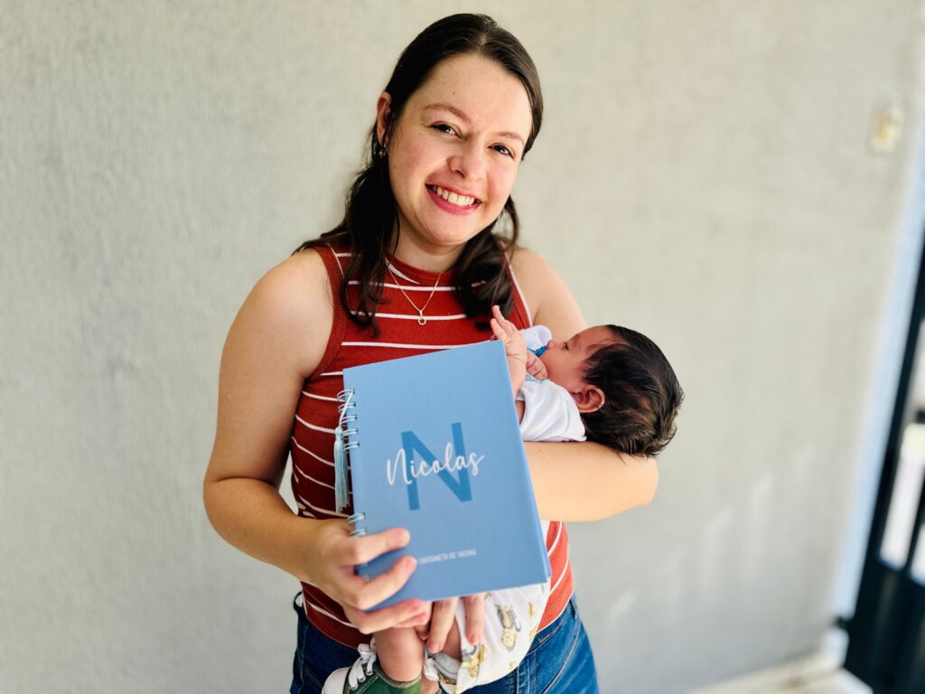 A mulher sorri para a câmera enquanto segura um bebê recém-nascido em seus braços. Ela está usando uma blusa listrada vermelha e branca e jeans azuis. O bebê está enrolado em uma manta com estampa de macaco e usa uma chupeta. A mulher segura um caderno azul com a letra "N" e o nome "Nicolas" escritos nele. O fundo é uma parede de concreto clara.