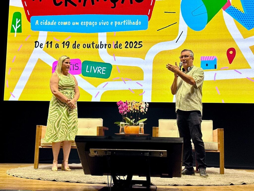 A imagem mostra um homem e uma mulher em um palco, possivelmente durante um evento ou apresentação. O homem, que está à direita, usa óculos, uma camisa xadrez de manga curta e calças escuras. Ele está de pé, segurando um microfone e gesticulando com as mãos enquanto fala. Há um fone de ouvido em seu pescoço. A mulher, à esquerda, veste um vestido estampado com um padrão de listras verdes e brancas. Ela está de pé com as mãos juntas na frente do corpo. Ambos estão em frente a um grande telão que exibe um fundo colorido com elementos gráficos que lembram um mapa. No telão, há textos em português: "A cidade como um espaço vivo e partilhado" e "De 11 a 19 de outubro de 2025". Há também botões estilizados com as palavras "IS" e "LIVRE". No palco, entre eles, há uma mesa baixa com um arranjo de flores cor-de-rosa e um vaso com líquido. Atrás deles, há cadeiras de madeira com almofadas claras. O palco está coberto por um tapete redondo e claro.