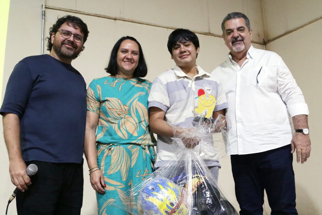 A imagem mostra quatro pessoas em pé, sorrindo para a câmera. Da esquerda para a direita, um homem com óculos e barba usa uma camiseta azul escura e calças pretas, segurando um microfone. Ao lado dele, uma mulher com cabelo escuro usa um vestido verde com estampa floral. Em seguida, um jovem com cabelo escuro veste uma camisa polo listrada e segura um troféu em forma de rosto sorridente amarelo com um chapéu vermelho, junto com uma cesta de presentes embrulhada em plástico transparente. Finalmente, um homem com cabelo grisalho e barba curta usa uma camisa branca e calça azul escura, com um relógio no pulso esquerdo. A iluminação sugere um evento interno, possivelmente uma premiação ou celebração.