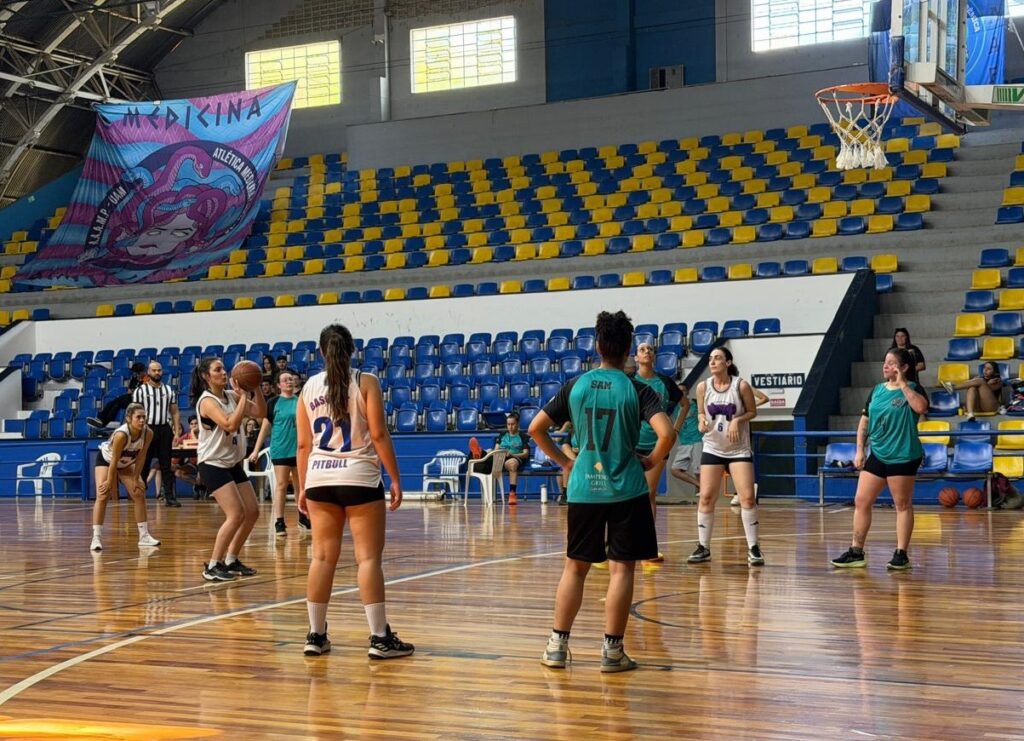 A imagem mostra uma partida de basquete feminino em uma quadra interna. Várias jogadoras estão em quadra, algumas em ação e outras aguardando. Uma jogadora de camisa branca com o número 21 está com a bola, prestes a arremessar. Outras jogadoras usam camisas verdes e brancas. Ao fundo, as arquibancadas estão parcialmente ocupadas por espectadores e há uma faixa grande com o nome "MEDICINA ATLETICA MEDICINA". O piso da quadra é de madeira polida e o aro da cesta de basquete é visível no canto superior direito.