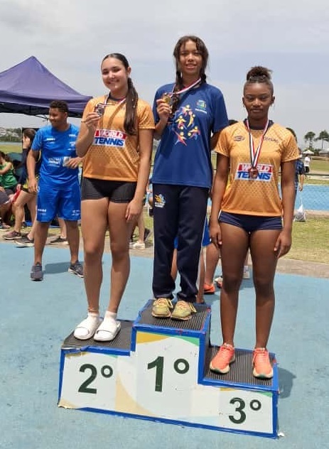 A imagem mostra três jovens mulheres em um pódio, celebrando suas conquistas em uma competição. Elas estão vestindo uniformes esportivos e segurando medalhas de ouro, prata e bronze. A mulher no centro, no primeiro lugar, usa um uniforme azul com um design colorido no peito e medalha de ouro. À sua esquerda, no segundo lugar, está uma mulher com um uniforme laranja com a inscrição "WORLD TENNIS" e uma medalha de ouro. À direita, no terceiro lugar, outra mulher com o mesmo uniforme laranja e medalha de ouro. O pódio está em um ambiente externo, possivelmente uma pista de atletismo, com uma tenda azul ao fundo e outras pessoas observando a cerimônia.