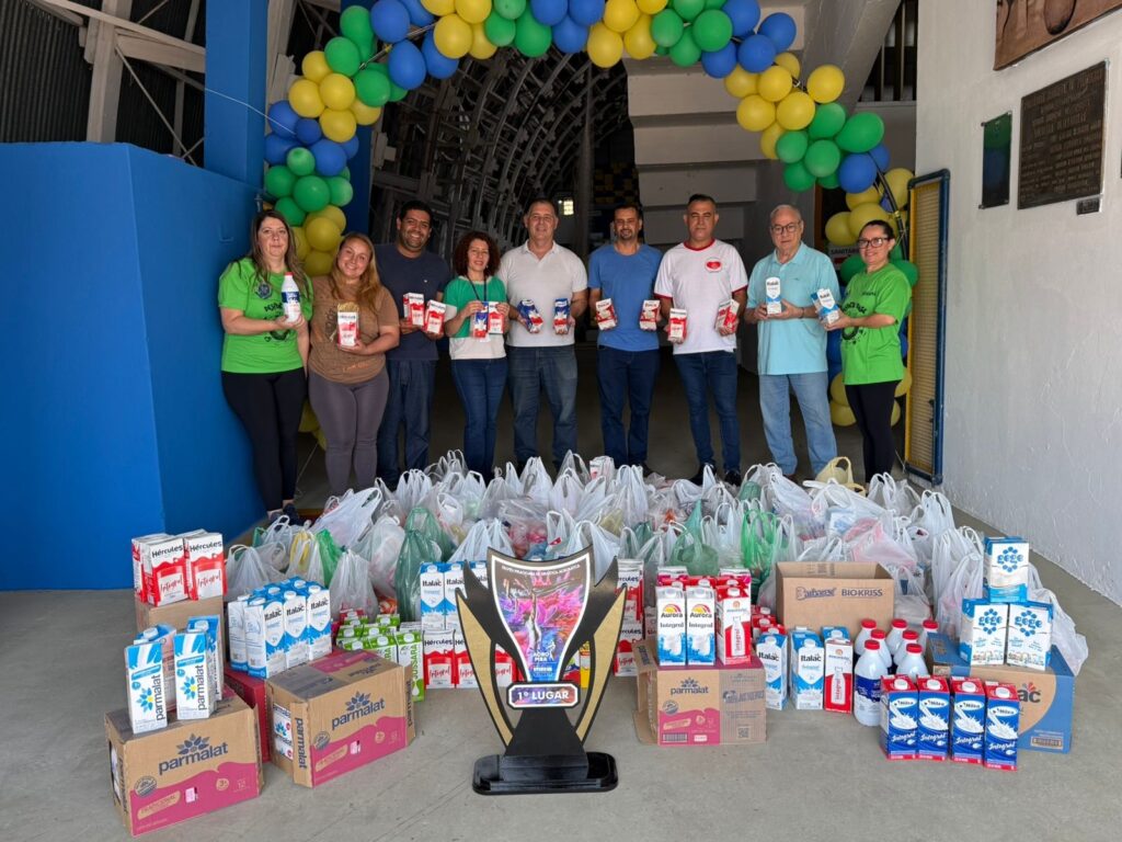A imagem mostra um grupo de 9 pessoas, 4 mulheres e 5 homens, em pé atrás de uma grande quantidade de sacolas plásticas cheias de alimentos e caixas de leite. Eles estão todos sorrindo e segurando caixas de leite ou produtos similares. Há um arco de balões nas cores verde, amarelo e azul acima deles. No centro da imagem, um troféu com a inscrição "1º LUGAR" está posicionado em cima de algumas caixas de leite. O cenário parece ser uma área externa coberta, possivelmente a entrada de um prédio ou um galpão. As pessoas vestem roupas casuais, e algumas usam camisetas com logotipos ou frases. As sacolas e caixas de produtos lácteos sugerem uma doação ou distribuição de alimentos.