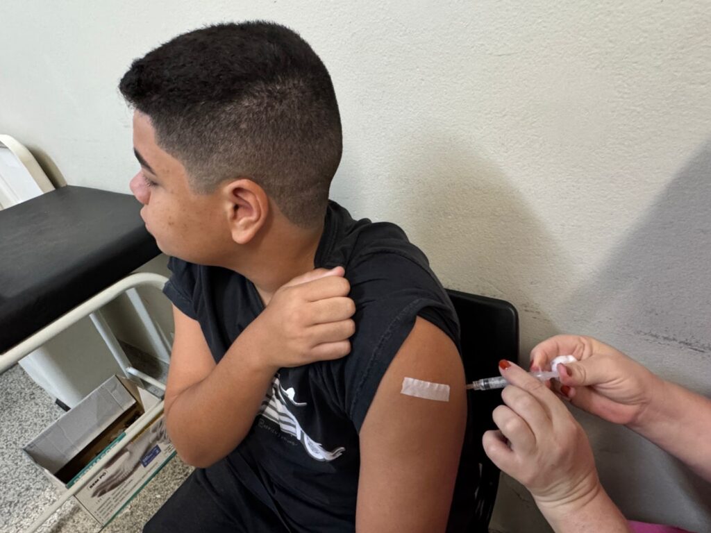 A imagem mostra um jovem de pele clara, com cabelo curto e escuro, vestindo uma camiseta preta. Ele está sentado, virado para a esquerda, com o braço direito levantado e o antebraço apoiado no ombro esquerdo. Uma bandagem branca está aplicada na parte superior do braço direito, onde uma agulha de injeção está sendo inserida. Uma pessoa, cujas mãos são visíveis, está aplicando a injeção, segurando a seringa e um algodão. O fundo é uma parede de cor clara, com uma sombra diagonal. À esquerda, parte de uma maca médica preta e uma caixa de papelão com algumas caixas dentro são visíveis.