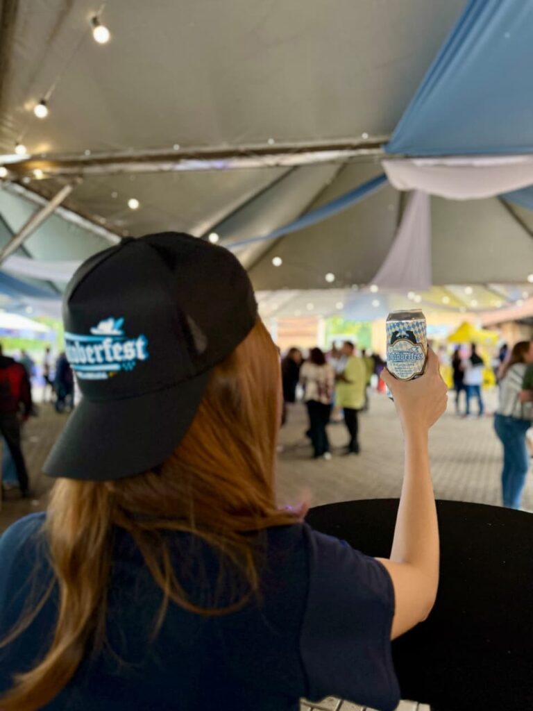 A mulher está usando um boné preto com o logo do Oktoberfest, e segurando uma lata de cerveja com a mão direita. Ela está em um evento, possivelmente uma festa ou festival, com outras pessoas ao fundo. O local parece ser ao ar livre, com tendas e luzes penduradas no teto.