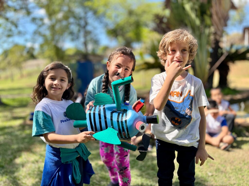 A imagem mostra três crianças sorrindo e segurando um peixe feito de material reciclado. A criança do lado esquerdo é uma menina com cabelos escuros ondulados, usando uma camiseta branca com detalhes verdes e uma jaqueta azul amarrada na cintura. Ao lado dela, uma menina com tranças longas, vestindo uma blusa verde e calças coloridas, segura o peixe com as duas mãos. A criança do lado direito é um menino com cabelos loiros e cacheados, usando uma camiseta branca com estampa do Batman e calças escuras. Ele está fazendo um gesto com a mão. O peixe é feito de garrafas plásticas azuis, com detalhes verdes e pretos, e parece ter sido decorado com outros materiais reciclados. As crianças estão ao ar livre, em um gramado com árvores e arbustos ao fundo, sugerindo um ambiente escolar ou de lazer. Há outras pessoas desfocadas ao fundo.