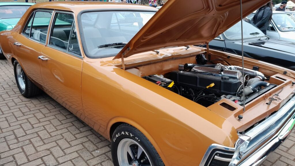 A imagem exibe um carro clássico de cor âmbar, com o capô aberto, revelando o motor. O carro está estacionado em um pátio pavimentado com tijolos. Ao fundo, outros carros e pessoas podem ser vistos, sugerindo um evento ou exposição de carros. O motor do carro âmbar parece ter sido modificado, com componentes cromados e tubulações visíveis. A pintura do carro é brilhante e reflete a luz ambiente. Os detalhes do carro, como as maçanetas cromadas e as rodas esportivas com pneus "GALAXY R1", são nítidos.