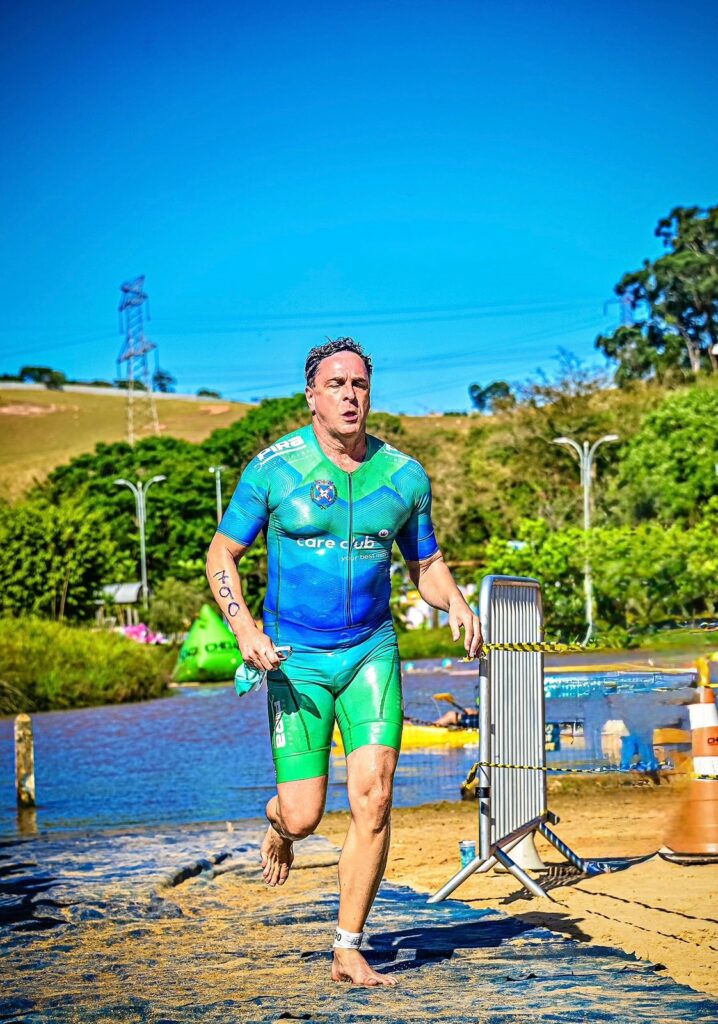 O atleta está correndo em terra firme, com uma paisagem natural ao fundo. Ele usa um traje de triatlo azul e verde, com o logo "care club" no peito. Seus braços e pernas estão marcados com números de competição, 760 no braço esquerdo e 30 no tornozelo direito. Ele corre descalço, com uma expressão de esforço no rosto, segurando uma peça de roupa na mão esquerda. Ao fundo, um lago azul e vegetação verde indicam um ambiente de prova ao ar livre, possivelmente um triatlo. O céu está claro e azul, com algumas nuvens esparsas.