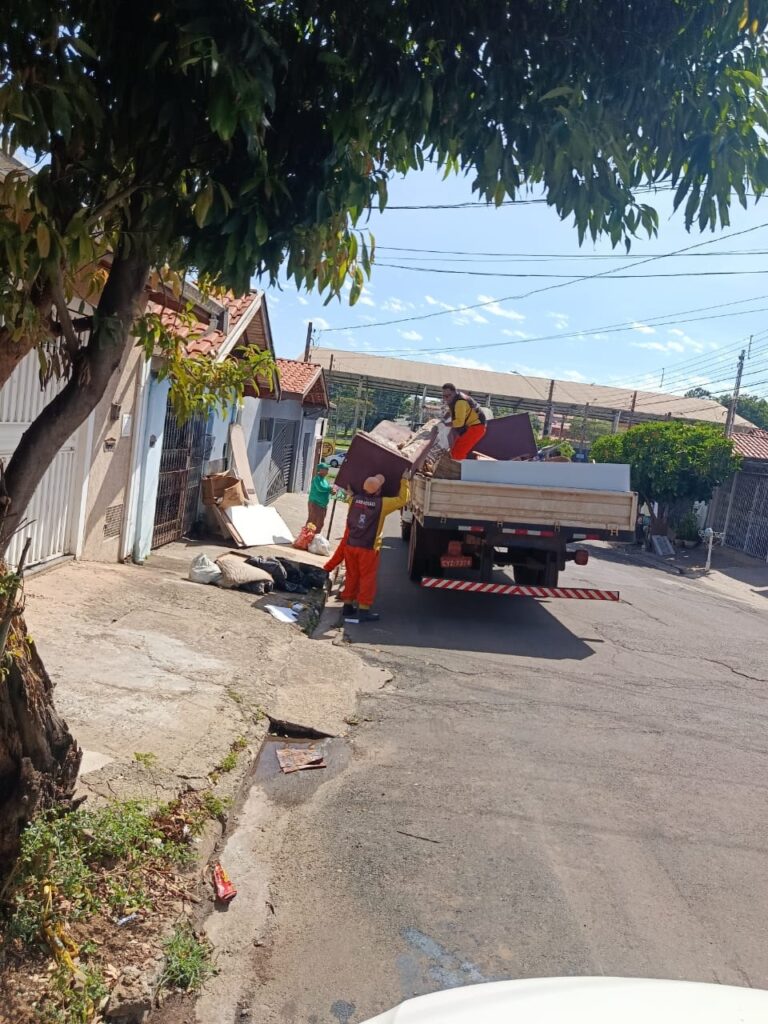 A imagem mostra dois trabalhadores vestindo uniformes de trabalho, um verde e outro laranja e amarelo, carregando móveis e outros itens para um caminhão de carga. O caminhão está estacionado na rua, com a parte traseira voltada para a câmera. Há uma casa à esquerda da imagem com uma árvore grande na frente. O céu está azul com algumas nuvens. Os trabalhadores parecem estar removendo móveis velhos ou danificados, como um sofá e um armário, que estão empilhados na plataforma do caminhão. Um dos trabalhadores está segurando um pedaço de madeira, enquanto o outro está posicionado no caminhão, ajudando a carregar os itens. A rua parece ser residencial, com calçadas e algumas plantas crescendo nas bordas. Há lixo e entulho acumulados na calçada perto da casa, sugerindo que uma limpeza ou remoção de resíduos está em andamento. A placa do caminhão é visível, com o código "CYZ-7374".