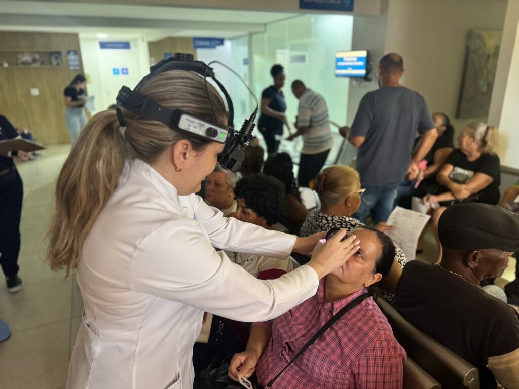 A imagem mostra uma médica oftalmologista examinando o olho de uma paciente em uma sala de espera. A médica está usando um oftalmoscópio de cabeça para iluminar e examinar o olho da paciente. A paciente está sentada, com a cabeça ligeiramente inclinada para trás, enquanto a médica segura delicadamente sua pálpebra para facilitar o exame. Ao fundo, outras pessoas aguardam em cadeiras, algumas lendo ou conversando. O ambiente parece ser uma clínica ou hospital, com sinalização e iluminação típicas desses locais. A cena transmite uma atmosfera de cuidado e atenção médica, com a profissional focada em seu exame e os pacientes em espera.