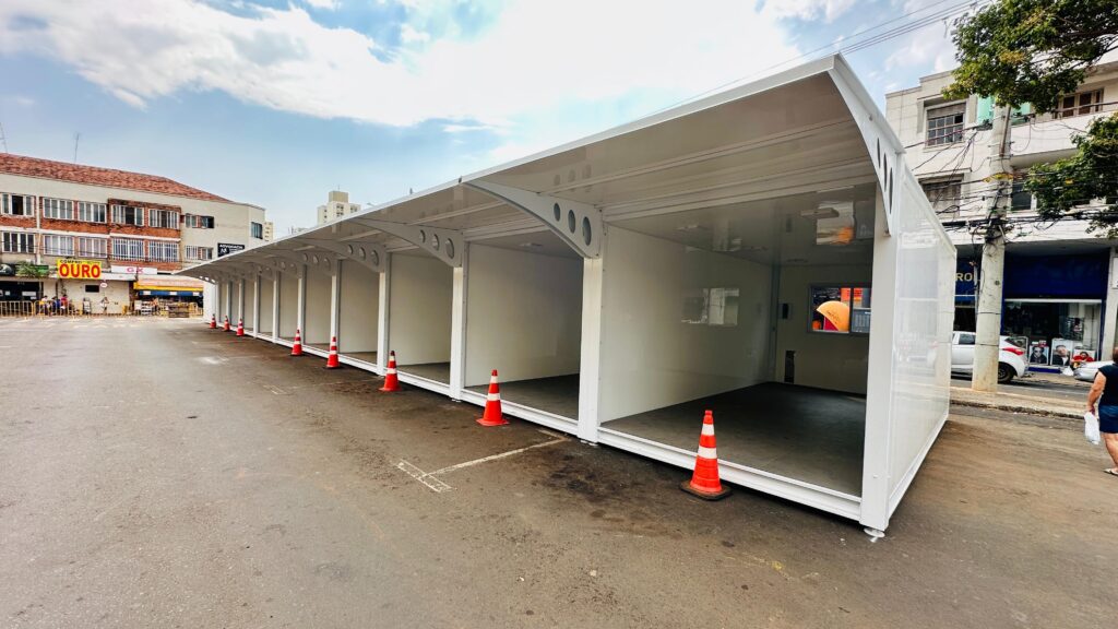 A imagem mostra uma estrutura modular branca, composta por várias unidades interligadas, que se estende ao longo de uma rua asfaltada. Cada unidade parece ser um pequeno compartimento com uma abertura frontal e um teto inclinado. Há cones de trânsito laranja e brancos posicionados em frente a algumas das unidades, sugerindo que a estrutura está em uma área pública, possivelmente uma rua ou estacionamento. Ao fundo, em um dos lados da rua, há edifícios com fachadas de tijolos e janelas, um deles com placas de estabelecimentos comerciais como "COMPRO OURO" e "GK". Do outro lado da estrutura modular, vemos mais edifícios, postes de luz com fiação aparente, e alguns carros estacionados. O céu está parcialmente nublado, com nuvens brancas e volumosas. Uma pessoa caminha na calçada ao fundo, perto de um dos edifícios. A iluminação sugere que é dia, com luz solar incidindo sobre a cena. A estrutura modular parece ser nova e ainda não está completamente ocupada ou decorada.