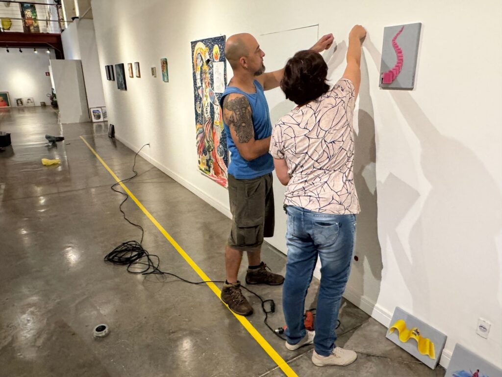 A imagem mostra duas pessoas, um homem e uma mulher, pendurando obras de arte em uma parede branca em uma galeria de arte. O homem, careca e com tatuagens no braço, veste uma regata azul e shorts cargo. A mulher, com cabelo escuro e curto, usa uma blusa estampada e jeans. Eles estão posicionando uma pequena pintura com uma forma abstrata rosa em zigue-zague. Ao lado da obra que estão pendurando, há outra pintura com um fundo azul escuro e uma figura colorida e detalhada, possivelmente religiosa ou mitológica. No chão, há cabos pretos enrolados e uma linha amarela pintada no concreto polido, que parece delimitar um caminho ou área de trabalho. Mais adiante na galeria, outras obras de arte estão expostas nas paredes, incluindo quadros menores e pinturas. O ambiente parece ser um espaço de exposição de arte com iluminação artificial.