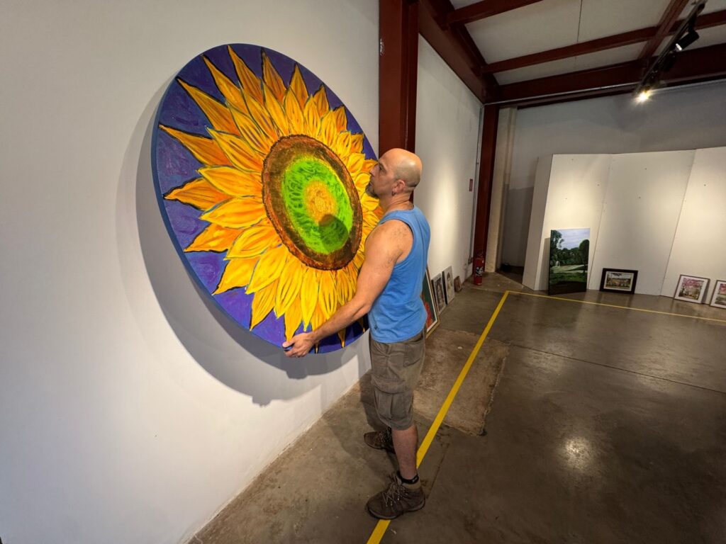 Um homem careca, vestindo uma regata azul e shorts cáqui, está em uma galeria de arte, ajustando uma grande pintura circular de um girassol. A pintura tem pétalas amarelas vibrantes e um centro marrom e verde, com um fundo roxo. O homem está de pé em um piso de concreto e há outras obras de arte expostas nas paredes brancas ao fundo. Uma linha amarela delimita uma área no chão da galeria.