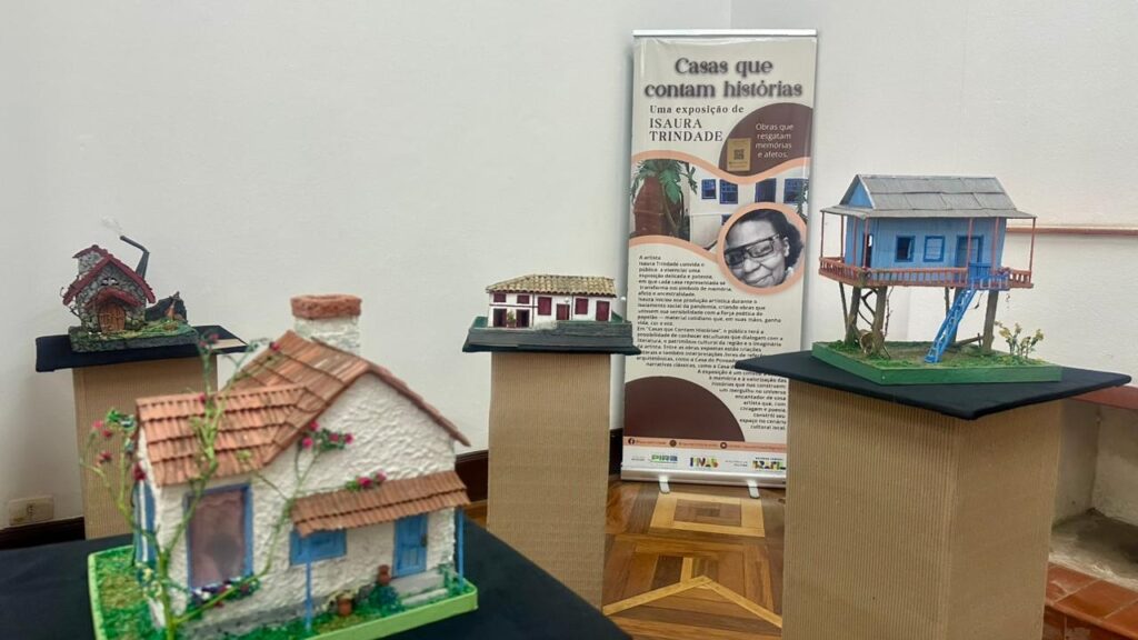 A imagem exibe uma exposição de maquetes de casas, cada uma com detalhes que sugerem histórias. No centro, um banner com o título "Casas que contam histórias" e a menção de uma exposição de Isaura Trindade. O banner também apresenta uma foto da artista e textos explicativos sobre a mostra. As maquetes apresentadas são variadas: * Uma casa com telhado de telhas vermelhas, paredes brancas e detalhes em azul, com uma pequena escada na frente e um jardim ao redor. * Uma casa menor, com telhado de pedra e chaminé, em um cenário mais rústico com vegetação. * Uma casa de dois andares, pintada de azul, elevada em palafitas, com uma escada azul que leva à varanda. As maquetes estão dispostas sobre pedestais de papelão, criando um ambiente de galeria. O fundo é uma parede branca, e o chão é de madeira com um padrão quadriculado. A iluminação destaca as obras, convidando o espectador a se aproximar e imaginar as narrativas que cada casa pode conter.