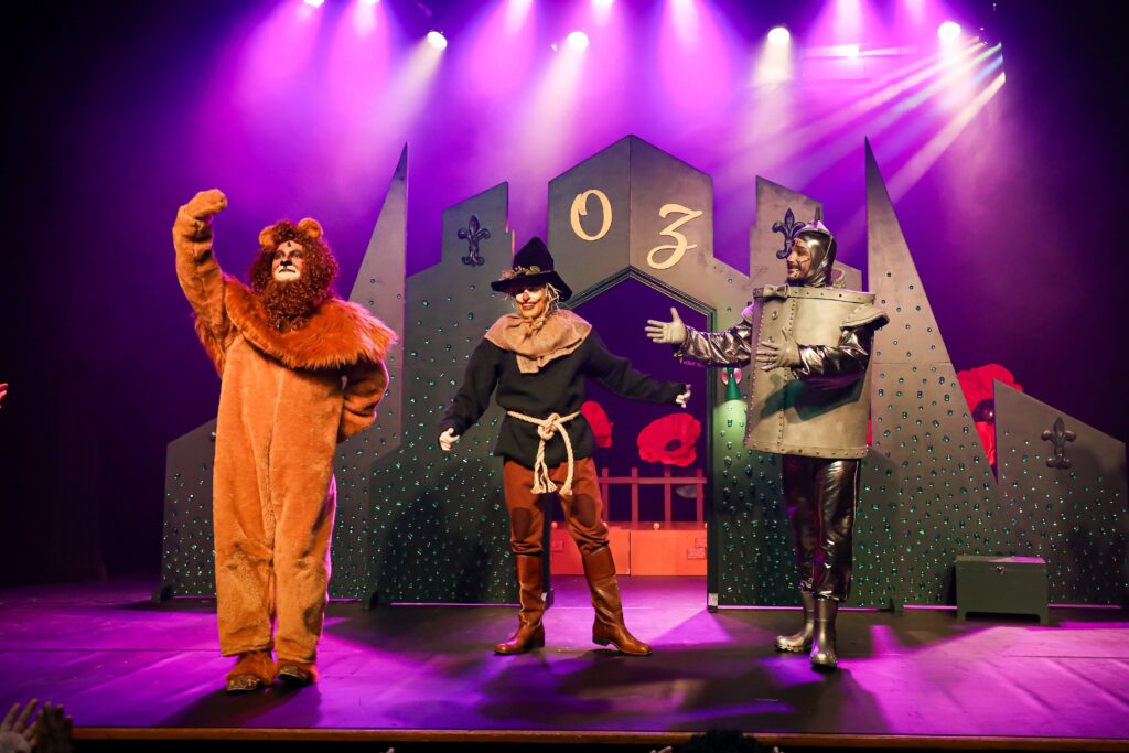 A imagem mostra uma cena de teatro com três personagens do "Mágico de Oz" no palco. Da esquerda para a direita, estão o Leão Covarde, o Espantalho e o Homem de Lata. Eles estão em frente a um cenário que representa a Cidade das Esmeraldas, com a palavra "OZ" em destaque. A iluminação é roxa e rosa, criando uma atmosfera mágica. O público, com as mãos levantadas, é visível na parte inferior da imagem, indicando que estão aplaudindo a performance.
