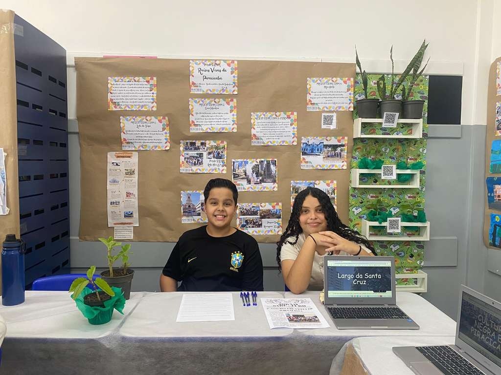 A imagem mostra uma exposição escolar com dois alunos, um menino e uma menina, sentados em uma mesa. Atrás deles, há um painel decorado com pôsteres, fotos e plantas em vasos. Os pôsteres parecem conter informações sobre a história ou cultura local, com títulos em português como "Raízes Vivas de Piracicaba". Há também QR codes espalhados pela exposição, provavelmente para acesso a mais informações. Um dos alunos está usando uma camiseta da seleção brasileira de futebol. Na mesa, há laptops abertos, papéis e canetas, sugerindo uma apresentação interativa. O ambiente parece ser uma feira de ciências ou um evento cultural em uma escola.