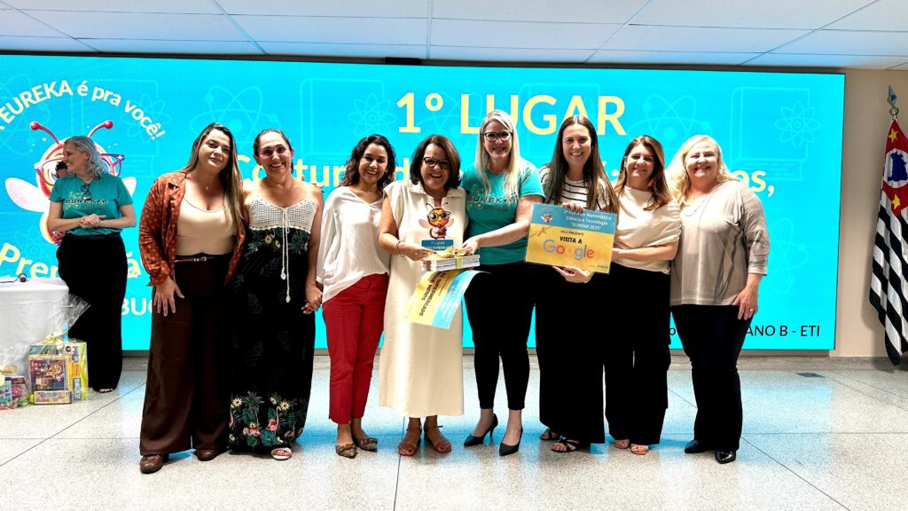 A imagem mostra um grupo de nove mulheres em pé em frente a um grande painel azul com a inscrição "EUREKA é pra você!" e "1° LUGAR". As mulheres parecem estar em um evento de premiação, possivelmente relacionado a ciência e tecnologia, dado o nome "EUREKA" e os símbolos científicos no painel. A maioria das mulheres está sorrindo e algumas seguram prêmios. A mulher no centro, usando um vestido branco, segura um monte de livros e um pequeno troféu com a imagem de uma abelha. A mulher à sua direita, usando uma camiseta verde, segura um certificado amarelo com o logo do Google e a inscrição "VALE PRESENTE VISITA A GOOGLE". Há também uma bandeira do estado de São Paulo à direita da imagem. As mulheres vestem roupas variadas, desde vestidos longos até calças e blusas. A iluminação sugere um evento interno. O painel ao fundo, com a mensagem em português, indica que o evento é no Brasil.