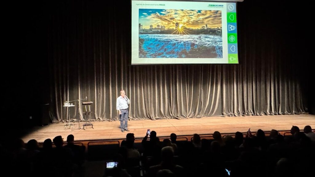 A imagem mostra um homem palestrando em um palco, com uma grande tela ao fundo exibindo uma paisagem com cachoeiras e um pôr do sol em uma cidade. O homem, vestindo camisa social e calça jeans, está em pé, segurando um microfone. O palco é escuro, com cortinas pesadas ao fundo. Na plateia, pessoas sentadas assistem à apresentação, algumas delas com celulares em mãos, possivelmente tirando fotos ou gravando. A iluminação foca no palestrante e na tela.