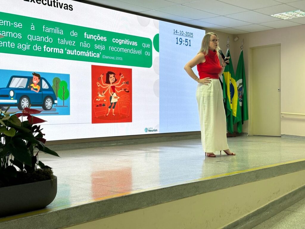 A imagem mostra uma mulher em pé em um palco, apresentando algo em uma tela grande. Ela está vestida com uma blusa vermelha de gola alta e calças brancas largas. Ela está segurando um microfone em uma mão e a outra mão está apoiada na cintura. A tela exibe texto em português e duas ilustrações. O texto fala sobre "funções cognitivas" e a ideia de agir de forma "automática". Uma ilustração mostra um homem dirigindo um carro azul, e a outra mostra uma mulher com vários braços cozinhando em uma cozinha. Há também uma data e hora exibidas na tela: 14-10-2025, 19:51. Ao fundo, há bandeiras do Brasil e do estado de São Paulo. Há também um vaso de plantas no canto inferior esquerdo da imagem. A iluminação sugere que a apresentação está acontecendo em um auditório ou sala de conferências.