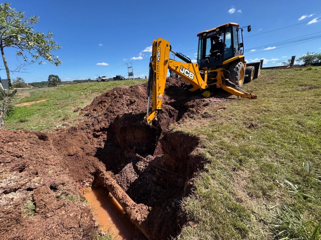 A imagem mostra uma escavadeira JCB amarela em um canteiro de obras, com o braço da máquina parcialmente dentro de uma vala recém-escavada. A terra removida forma uma pilha de terra marrom ao lado da vala. Dentro da vala, um cano enferrujado e parcialmente submerso em água lamacenta é visível. O céu está azul claro com algumas nuvens brancas esparsas, sugerindo um dia ensolarado. Ao fundo, pode-se ver uma paisagem com algumas árvores e postes de energia elétrica. Há também alguns veículos estacionados ao longe, indicando a presença de mais atividades na área. A grama verde ao redor da vala está bem aparada, contrastando com a terra exposta. O operador da escavadeira está visível dentro da cabine, concentrado em seu trabalho.