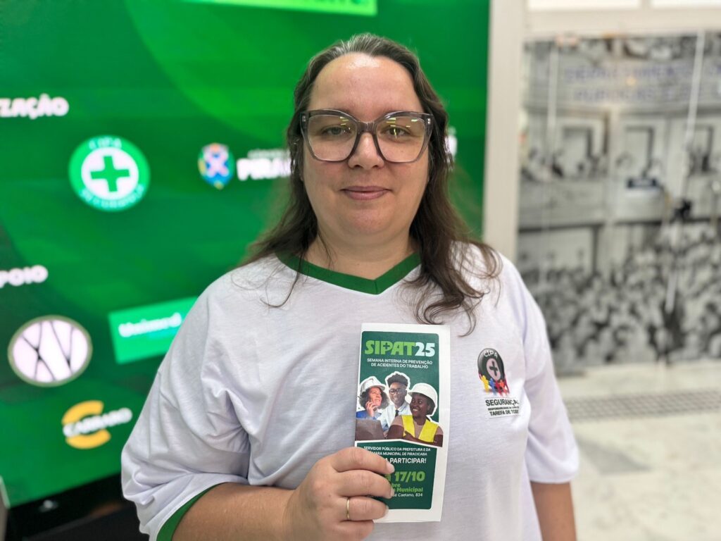 A imagem exibe uma mulher usando óculos e uma camiseta branca com detalhes verdes. Ela está segurando um folheto com informações sobre a "SIPAT25 - Semana Interna de Prevenção de Acidentes do Trabalho". Ao fundo, há um painel verde com logos e textos, incluindo um símbolo de cruz verde, e uma imagem em preto e branco de pessoas. A mulher sorri para a câmera, parecendo participar de um evento relacionado à segurança no trabalho.