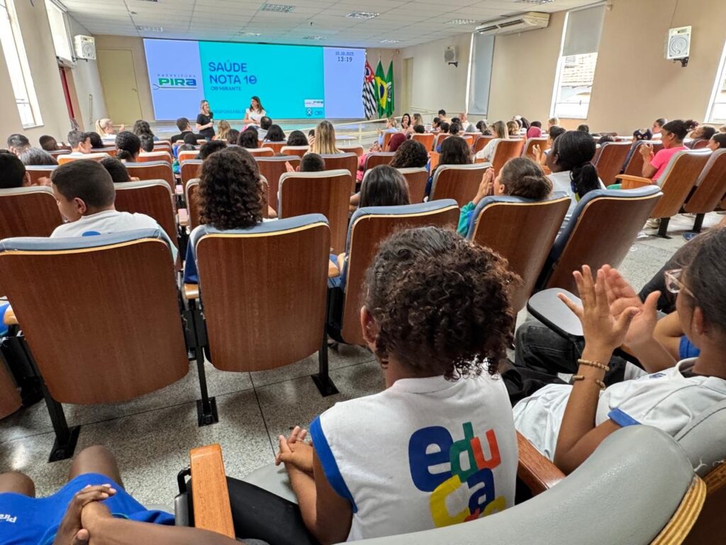 A imagem mostra uma sala de aula ou auditório, onde um grupo de estudantes, em sua maioria crianças e adolescentes, está sentado em cadeiras de madeira com estofado azul. No centro da sala, no palco, duas mulheres estão apresentando algo em uma tela grande. A tela exibe o título "SAÚDE NOTA 10" e o nome "PIRA O MIRANTE", juntamente com a data "10.10.2023" e o horário "13:19". Há também bandeiras do estado de São Paulo e do Brasil ao lado da tela. Os estudantes parecem atentos à apresentação, alguns estão com as mãos levantadas como se fossem fazer perguntas ou aplaudir. As cadeiras estão dispostas em fileiras, e a imagem foca principalmente na parte de trás das cabeças dos alunos, com alguns rostos visíveis nas primeiras fileiras. A iluminação na sala é clara, vindo de cima e das janelas, e o ambiente parece ser de um evento educacional ou informativo.