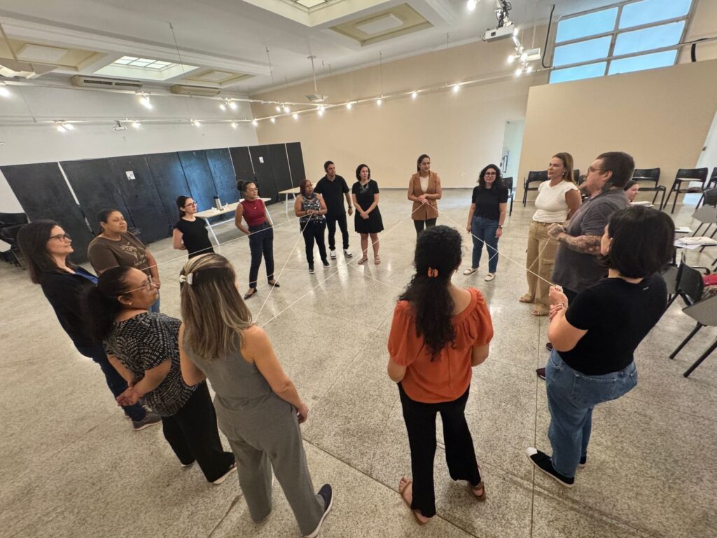 A imagem mostra um grupo de cerca de 11 pessoas em pé em um círculo em uma sala com piso de concreto e teto com iluminação embutida e luminárias. Várias linhas de barbante branco formam uma teia entre os participantes, que estão segurando o barbante em diferentes pontos. As pessoas estão vestidas casualmente, com algumas usando roupas de trabalho. Elas parecem estar participando de uma atividade de grupo ou workshop. A atmosfera parece ser colaborativa e engajadora. A sala parece ser um espaço multifuncional, possivelmente uma sala de aula ou um estúdio de arte, com algumas mesas e cadeiras nas laterais. Há também uma grande área escura com painéis na parede do fundo.