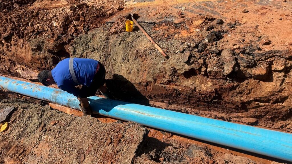 A imagem mostra um trabalhador agachado ao lado de um grande tubo azul que está parcialmente enterrado em uma vala. O chão ao redor da vala é composto de terra marrom e pedras, com alguns pedaços de asfalto visíveis na parte superior. Há uma pá e um balde amarelo próximos à vala, sugerindo que o trabalho de escavação está em andamento. O trabalhador, vestindo uma camisa azul e calças escuras, parece estar inspecionando ou ajustando o tubo. A luz do sol ilumina a cena, criando sombras nítidas.