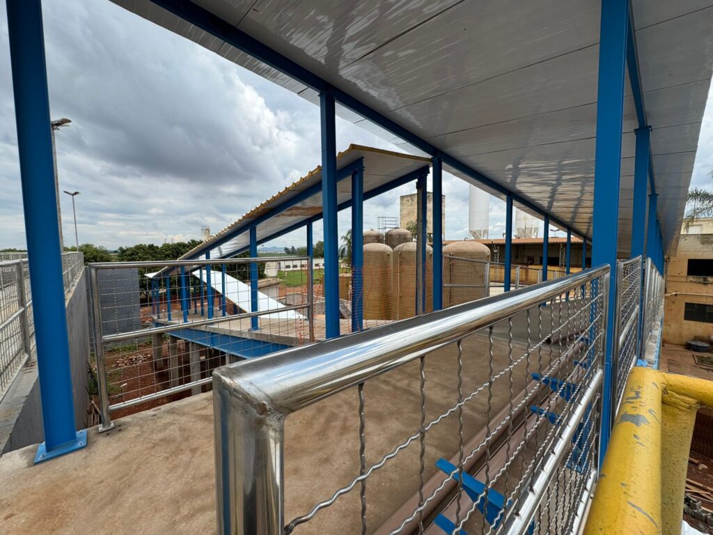 A imagem apresenta uma passarela elevada com estrutura metálica azul e cobertura de telhas metálicas. O piso da passarela é de concreto, e as laterais são protegidas por grades de metal com malha quadriculada e corrimãos de aço inoxidável polido. Ao fundo, observa-se um complexo industrial com vários tanques de armazenamento de grande porte, alguns de cor bege e outros brancos. Há também edificações com características industriais, vegetação e um céu nublado. A passarela parece conectar diferentes áreas desse complexo, oferecendo um caminho seguro e coberto.