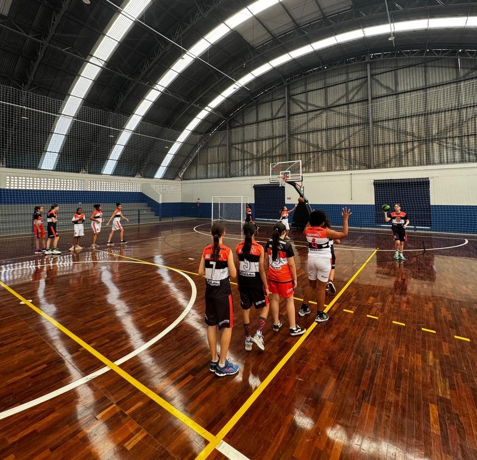 A imagem mostra um grupo de jovens jogadoras de basquete em uma quadra interna. O piso da quadra é de madeira brilhante, com linhas amarelas e brancas demarcando as áreas de jogo. Ao fundo, uma estrutura metálica com telhado curvo e janelas permite a entrada de luz natural. As jogadoras, vestindo uniformes de basquete em tons de preto, laranja e branco, estão espalhadas pela quadra. Algumas estão em formação, enquanto outras parecem estar em meio a uma jogada ou treinamento. Uma jogadora no lado direito da quadra está com a bola na mão, pronta para arremessar, e outra, mais ao fundo, parece estar defendendo. A atmosfera geral sugere um ambiente de treino ou jogo competitivo.
