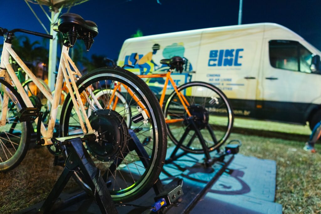 A imagem mostra duas bicicletas em um suporte, prontas para uso. A bicicleta em primeiro plano é branca e está posicionada à esquerda, enquanto uma bicicleta laranja está ao fundo, à direita. Ambas as bicicletas parecem ser elétricas, com motores visíveis nos cubos das rodas traseiras. Ao fundo, um furgão branco com o logotipo "BIKE" em letras azuis estilizadas está estacionado. O furgão tem um design gráfico com figuras de pessoas andando de bicicleta. A cena noturna, com iluminação artificial, sugere um evento ou demonstração relacionado a bicicletas.