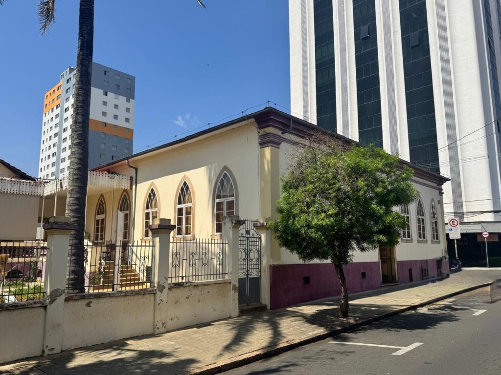 A imagem mostra uma rua em uma cidade com vários prédios. No primeiro plano, há um prédio baixo e antigo, com uma fachada amarela e detalhes em roxo. Ele tem janelas em arco e uma cerca de ferro na frente. Uma árvore pequena e frondosa cresce em frente a este prédio. Ao lado, erguem-se dois prédios modernos e altos. Um deles tem uma fachada listrada em tons de cinza e branco, com muitas janelas. O outro prédio, mais à esquerda, é mais colorido, com faixas horizontais em tons de laranja, cinza e branco. O céu está azul e limpo, indicando um dia ensolarado. A rua tem calçada e sinalização de trânsito visível.