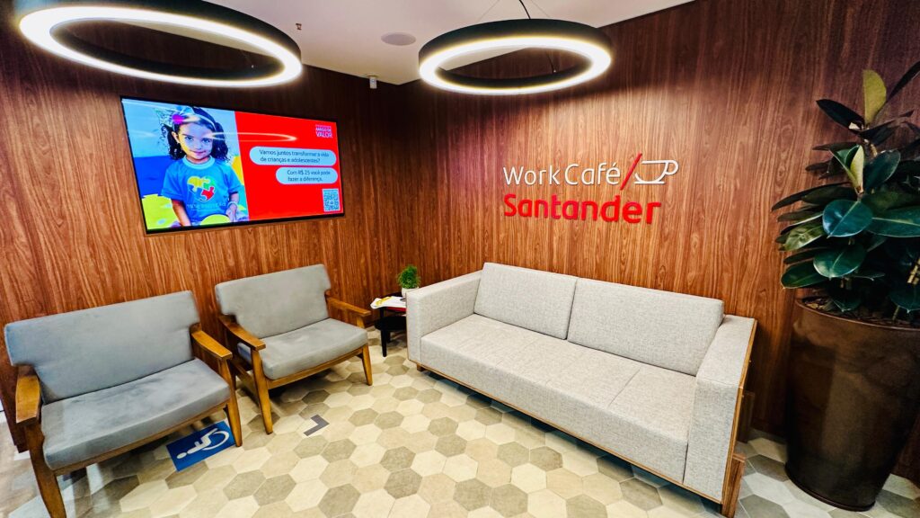 A imagem retrata um espaço de convivência com um design moderno e acolhedor, possivelmente um "Work Café" do Santander. As paredes são revestidas em madeira com um padrão vertical, criando uma atmosfera quente e sofisticada. No canto esquerdo, uma tela exibe uma imagem vibrante com a mensagem "Amigo do Valor" e um apelo para transformar a vida de crianças e adolescentes, incentivando doações a partir de R$ 25. Ao lado da tela, duas poltronas cinzas com estrutura de madeira oferecem um local para sentar. Uma delas tem um adesivo azul no chão indicando acessibilidade para pessoas com deficiência. À direita, um sofá cinza de três lugares convida ao relaxamento. Acima do sofá, em destaque na parede de madeira, está a logo "Work Café Santander" em letras vermelhas e um ícone estilizado de xícara de café. A iluminação é proporcionada por dois grandes anéis de luz suspensos no teto, que emitem uma luz branca e suave. O chão é revestido por um mosaico hexagonal em tons de bege e cinza, adicionando um toque contemporâneo ao ambiente. Um grande vaso com uma planta exuberante, à direita do sofá, complementa a decoração, trazendo um elemento natural para o espaço.
