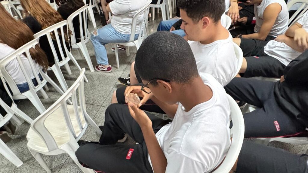 A imagem mostra um grupo de jovens sentados em cadeiras de plástico brancas, aparentemente em uma sala ou auditório. No centro da imagem, um jovem com óculos e uma camiseta branca está concentrado em examinar um objeto transparente que segura nas mãos. Ao seu redor, outros jovens também vestem camisetas brancas e calças pretas, alguns com detalhes vermelhos. O ambiente parece ser informal, com os jovens em posições relaxadas, alguns conversando e outros observando o que acontece. O foco principal recai sobre o jovem no centro, que demonstra interesse em analisar o objeto em suas mãos.
