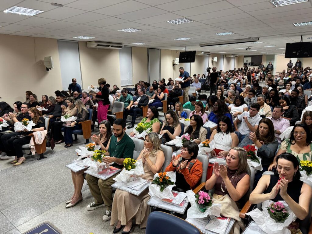 Um grande grupo de pessoas está sentado em um auditório bem iluminado, participando de um evento formal. Muitas delas vestem roupas sociais e seguram pequenos arranjos de flores, sugerindo uma cerimônia de formatura, homenagem ou reconhecimento. As primeiras fileiras estão ocupadas por pessoas que parecem ser o foco da celebração, com flores e papéis ou certificados no colo. Algumas pessoas estão aplaudindo, indicando um momento de celebração. O ambiente tem telas grandes nas paredes, aparelhos de ar-condicionado e teto com iluminação embutida.